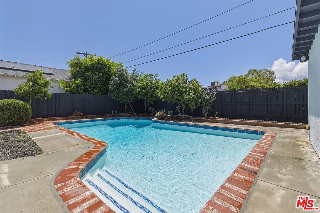 Backyard pool in daylight with brick coping, dark wood fence, and mature green shrubs along the perimeter